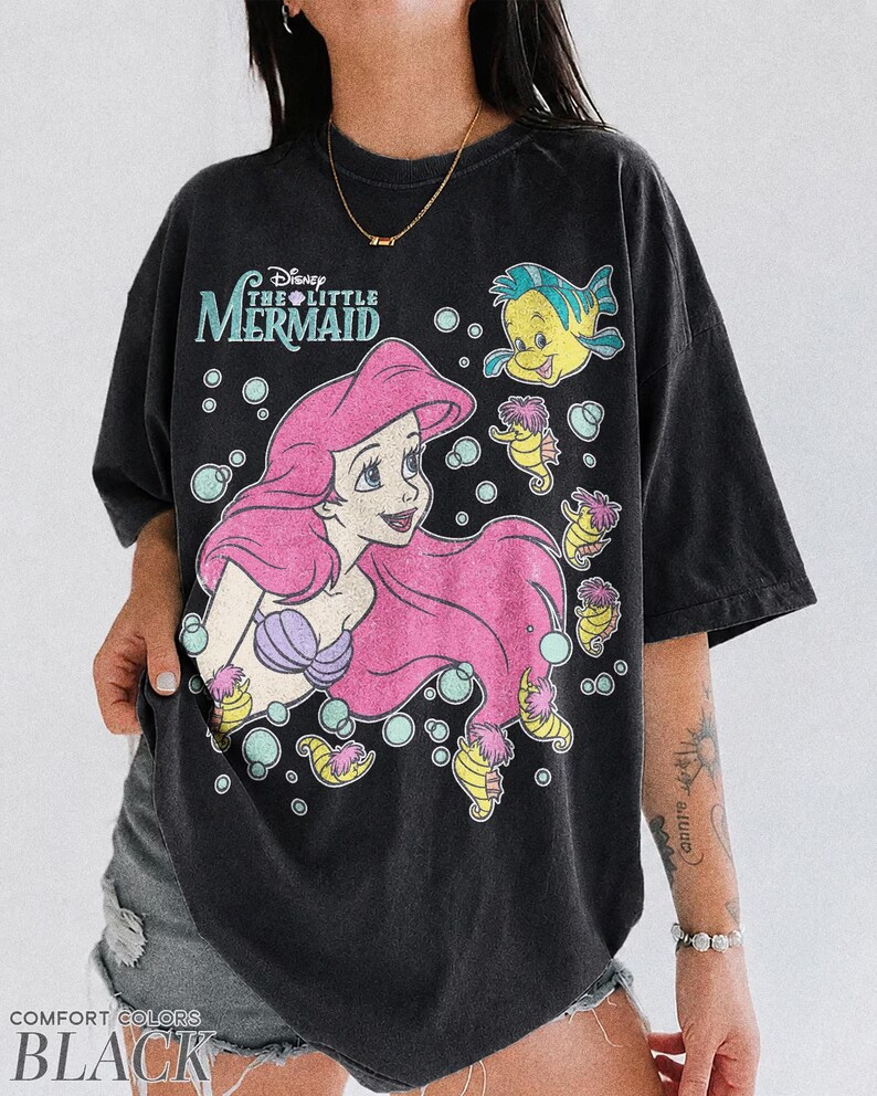 May include: A black short-sleeve t-shirt featuring a graphic print of Ariel from The Little Mermaid with pink hair, a purple seashell top, and Flounder. The design includes bubbles and small seahorses, with the text Disney The Little Mermaid.
