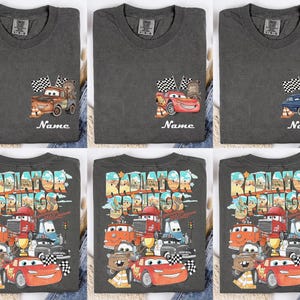 May include: Dark gray t-shirts featuring Cars movie characters. The shirts display images of Mater, Lightning McQueen, and other characters with the text "Name" below. The bottom row of shirts has a large "Radiator Springs" graphic.