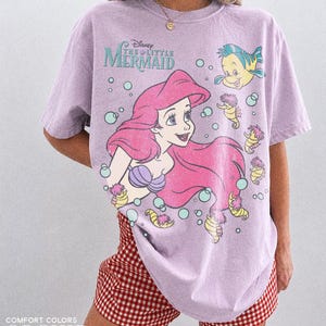 May include: A light purple short-sleeve t-shirt featuring a graphic of Ariel from Disney's The Little Mermaid, along with Flounder and several seahorses surrounded by bubbles. The shirt has a distressed print and the text Disney The Little Mermaid.