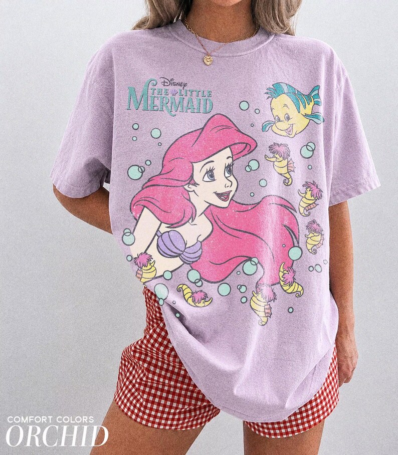 May include: A light purple short-sleeve t-shirt featuring a graphic of Ariel from Disney's The Little Mermaid, along with Flounder and several seahorses surrounded by bubbles. The shirt has a distressed print and the text Disney The Little Mermaid.