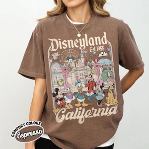 May include: Espresso-colored t-shirt featuring a Disneyland graphic. The design includes Mickey Mouse, Minnie Mouse, Donald Duck, Goofy, and Pluto in front of a park scene. The text "Disneyland Est. 1955 California" is also displayed.