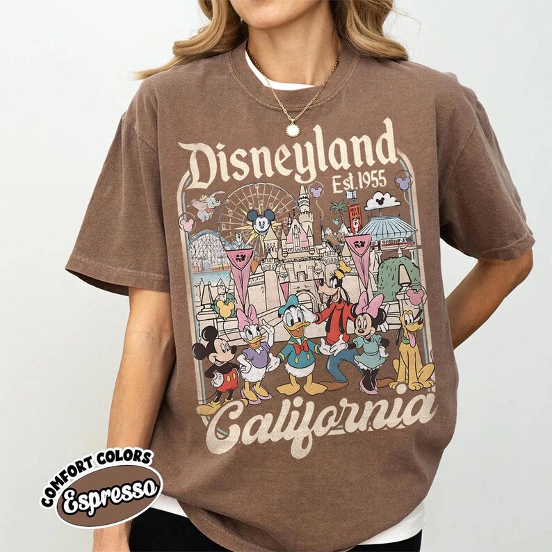 May include: Espresso-colored t-shirt featuring a Disneyland graphic. The design includes Mickey Mouse, Minnie Mouse, Donald Duck, Goofy, and Pluto in front of a park scene. The text "Disneyland Est. 1955 California" is also displayed.