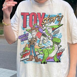 May include: Cream-colored t-shirt featuring a graphic of Woody, Buzz Lightyear, and Rex from Toy Story. The design includes the words "TOY Story" in red and gold, "Since 1995," and character names. The shirt has a classic, vintage style.