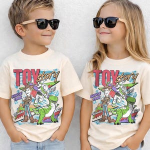 May include: Two children wearing cream-colored t-shirts with a "Toy Story" graphic. The graphic features Woody, Buzz Lightyear, and Rex, with the text "TOY Story" and "SINCE 1995". Both children are wearing black sunglasses.
