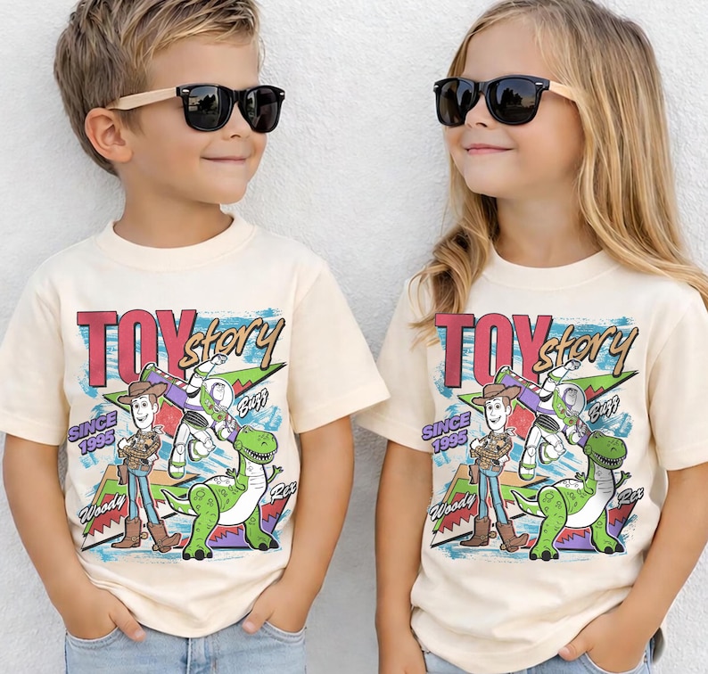 May include: Two children wearing cream-colored t-shirts with a "Toy Story" graphic. The graphic features Woody, Buzz Lightyear, and Rex, with the text "TOY Story" and "SINCE 1995". Both children are wearing black sunglasses.