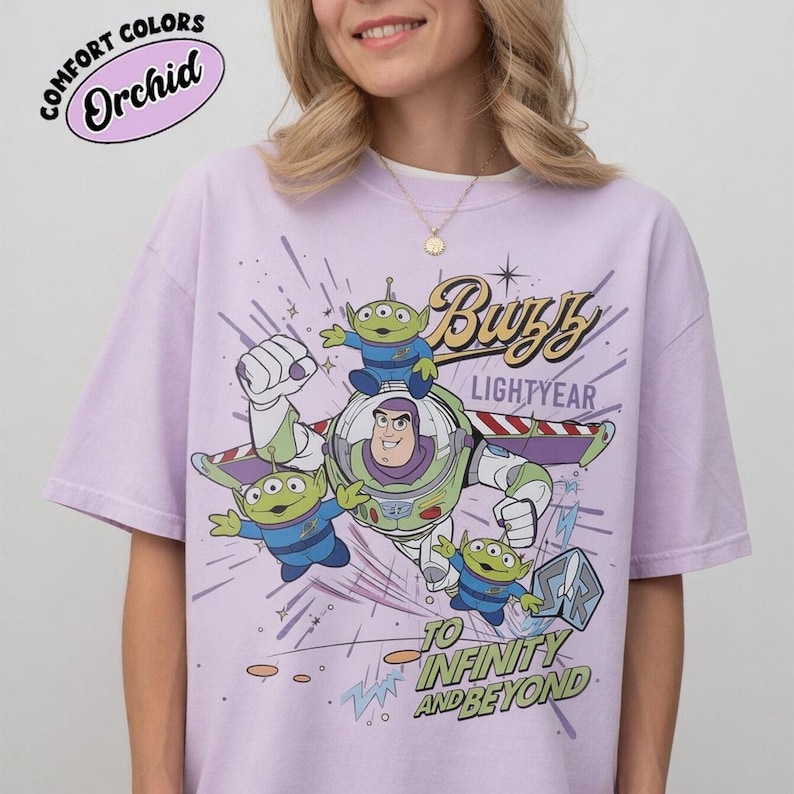 May include: A light purple t-shirt featuring a graphic of Buzz Lightyear and the Little Green Men from Toy Story. The text "Buzz Lightyear" and "To Infinity and Beyond" are also displayed. The shirt has short sleeves and a crew neck.