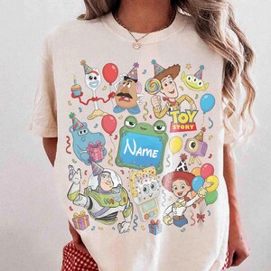 May include: A cream-colored t-shirt featuring a colorful graphic print of various Toy Story characters wearing party hats, including Woody, Buzz Lightyear, and Jessie, surrounded by balloons, gifts, and a central green frame with the text Name.