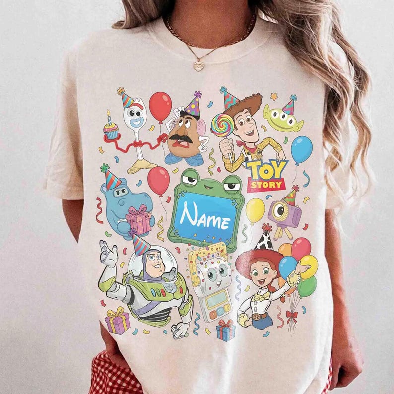 May include: A cream-colored t-shirt featuring a colorful graphic print of various Toy Story characters wearing party hats, including Woody, Buzz Lightyear, and Jessie, surrounded by balloons, gifts, and a central green frame with the text Name.