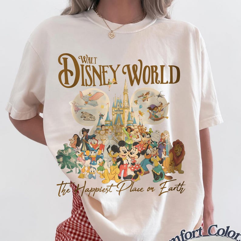 May include: Cream-colored t-shirt featuring a graphic of Disney characters and the text "Walt Disney World" in gold. The shirt includes images of Cinderella's castle, Mickey Mouse, and other beloved characters. The text "The Happiest Place on Earth" is also printed on the shirt.