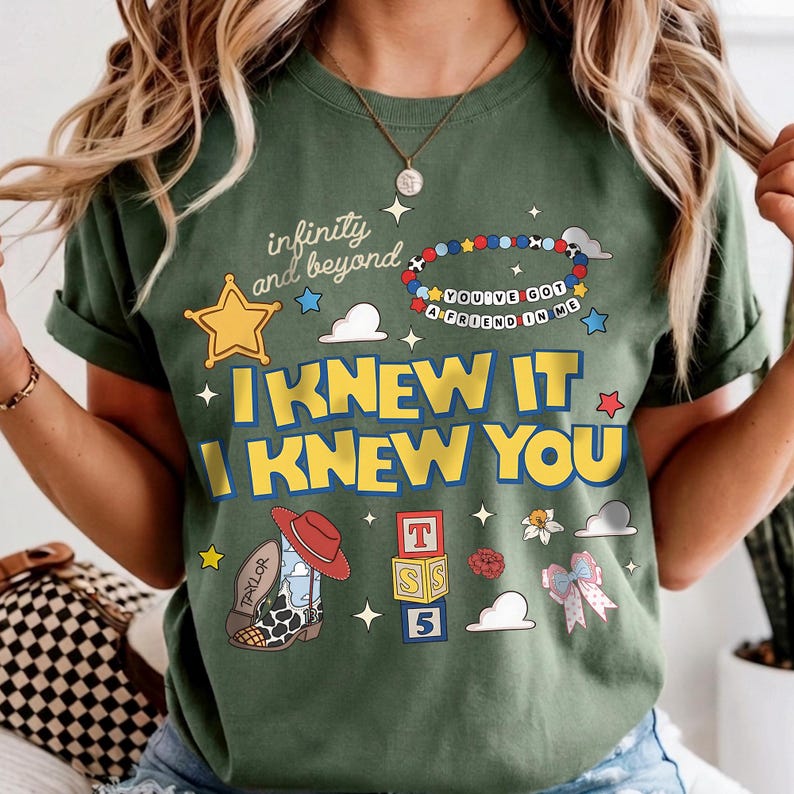 May include: An olive green t-shirt featuring a graphic print with the text "infinity and beyond", "YOU'VE GOT A FRIEND IN ME", "I KNEW IT", and "I KNEW YOU". The design includes a yellow star, a beaded bracelet, a cowboy boot, blocks, and a bow.