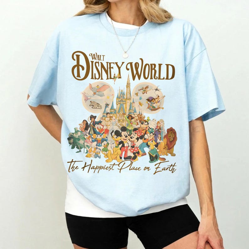 May include: Light blue t-shirt featuring the text "Walt Disney World" in a brown script above a colorful graphic of Disney characters and a castle. The phrase "the Happiest Place on Earth" is printed below the graphic.