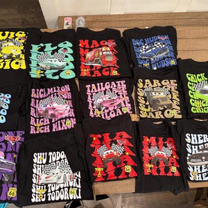 May include: A selection of black t-shirts showcasing characters and designs from the film "Cars." Each shirt features a different character with text, including "Luigi," "Guido," and "Sheriff." The shirts are displayed on a wooden surface.