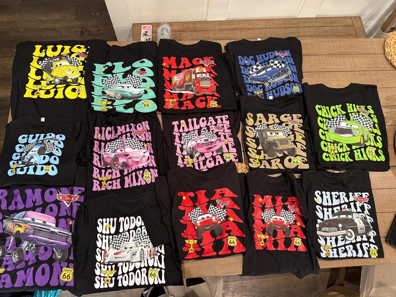 May include: A selection of black t-shirts showcasing characters and designs from the film "Cars." Each shirt features a different character with text, including "Luigi," "Guido," and "Sheriff." The shirts are displayed on a wooden surface.