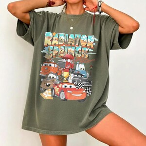 May include: Olive green t-shirt featuring a colorful graphic of characters from the movie "Cars" with the text "Radiator Springs". The design includes Lightning McQueen and other vehicles. The shirt has a relaxed fit.