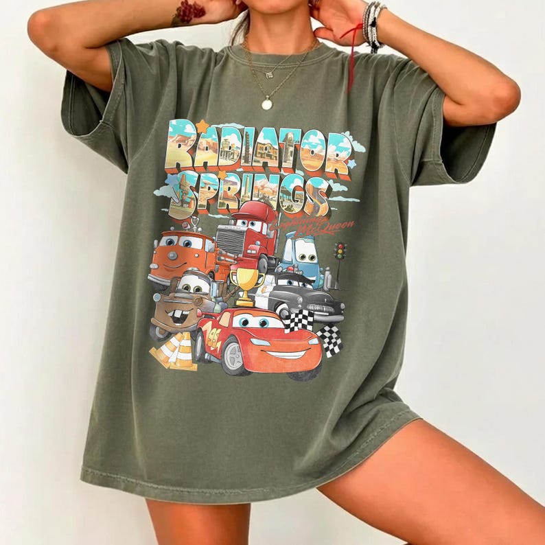 May include: Olive green t-shirt featuring a colorful graphic of characters from the movie "Cars" with the text "Radiator Springs". The design includes Lightning McQueen and other vehicles. The shirt has a relaxed fit.
