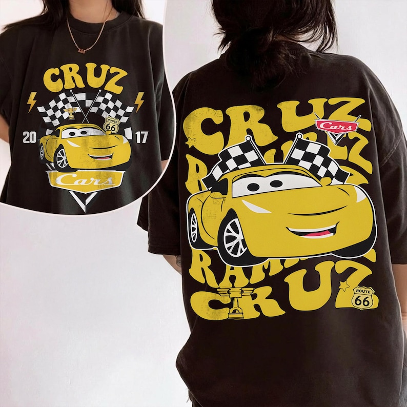May include: Black t-shirt with a yellow cartoon car design, including the text "CRUZ" and "Cars". The graphic features checkered flags, lightning bolts, and the number "66". The shirt has a vintage, worn appearance.