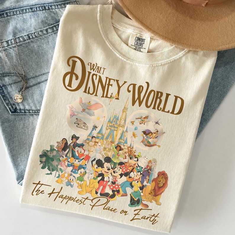 May include: Cream-colored t-shirt featuring the text "Walt Disney World" in brown script, with a graphic of Disney characters and Cinderella's castle. The phrase "The Happiest Place on Earth" is printed below the graphic.