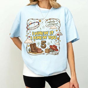 May include: A light blue t-shirt featuring a graphic print with the text "I KNEW IT I KNEW YOU", "5", and "Cinfinity and beyond". The design includes illustrations of cowboy boots, a red hat, alphabet blocks, stars, clouds, and a lasso rope border.