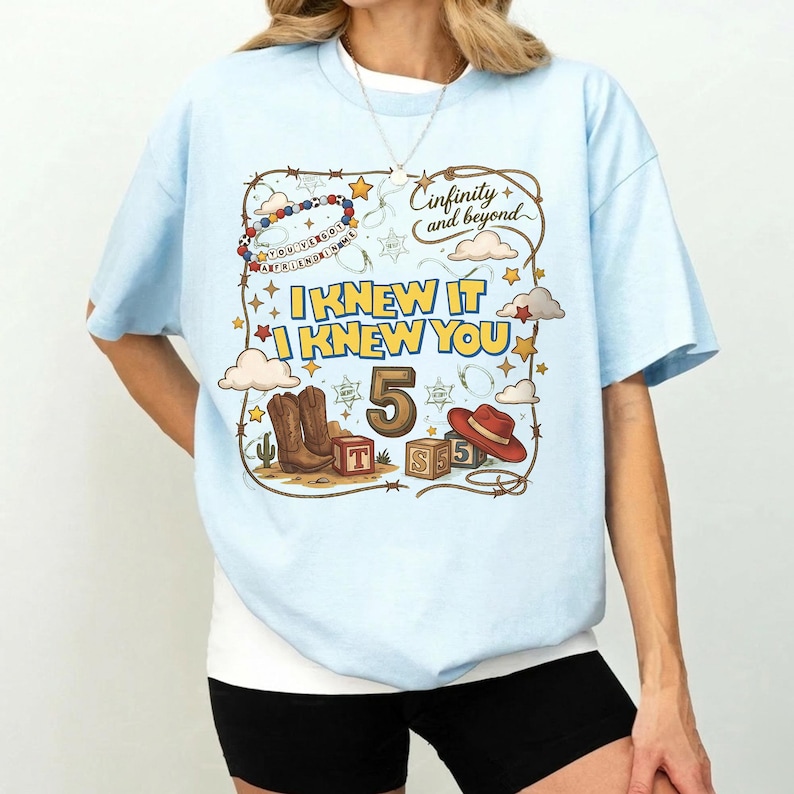 May include: A light blue t-shirt featuring a graphic print with the text "I KNEW IT I KNEW YOU", "5", and "Cinfinity and beyond". The design includes illustrations of cowboy boots, a red hat, alphabet blocks, stars, clouds, and a lasso rope border.