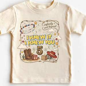 May include: A cream-colored t-shirt featuring a graphic print with the text "I KNEW IT I KNEW YOU", "5", and "Cinfinity and beyond". The design includes a beaded bracelet, cowboy boots, a red hat, alphabet blocks, stars, clouds, and a lasso.