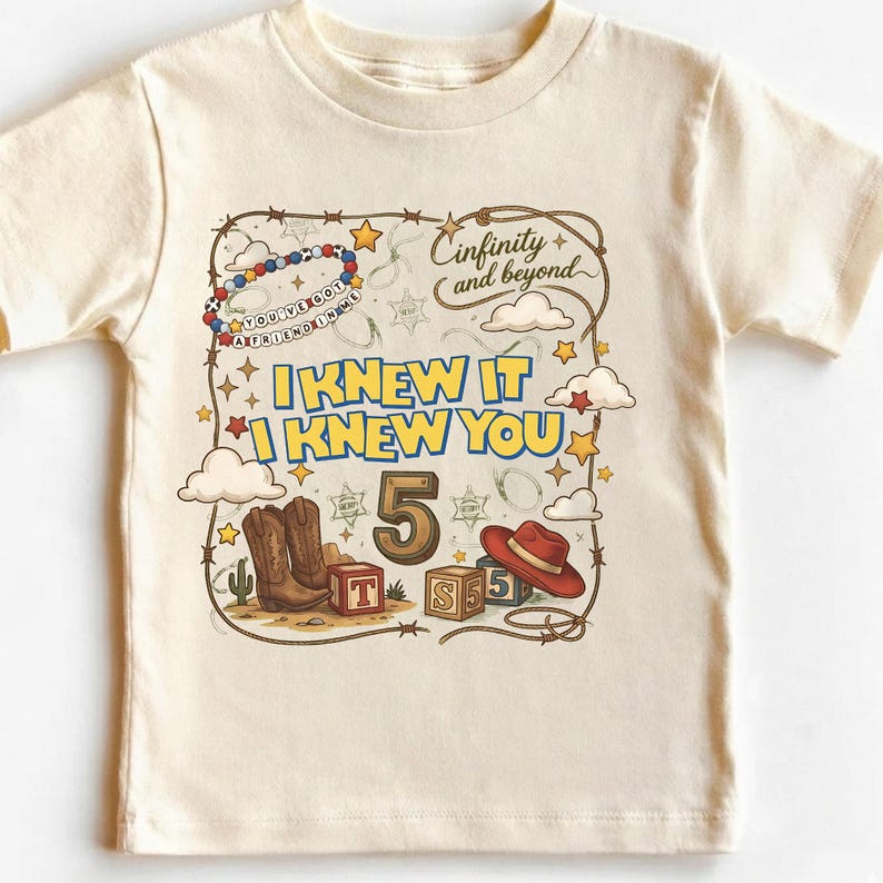May include: A cream-colored t-shirt featuring a graphic print with the text "I KNEW IT I KNEW YOU", "5", and "Cinfinity and beyond". The design includes a beaded bracelet, cowboy boots, a red hat, alphabet blocks, stars, clouds, and a lasso.