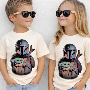 K&ouml;nnte beinhalten: Zwei cremefarbene Kurzarm-T-Shirts mit einem Grafikdruck eines behelmten Kriegers in dunkler R&uuml;stung, der ein kleines gr&uuml;nes Wesen mit gro&szlig;en Ohren h&auml;lt. Die T-Shirts werden mit schwarzen Sonnenbrillen mit hellen Holzrahmen getragen.