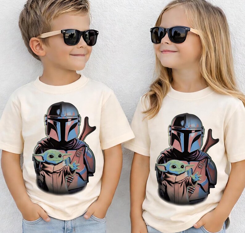 K&ouml;nnte beinhalten: Zwei cremefarbene Kurzarm-T-Shirts mit einem Grafikdruck eines behelmten Kriegers in dunkler R&uuml;stung, der ein kleines gr&uuml;nes Wesen mit gro&szlig;en Ohren h&auml;lt. Die T-Shirts werden mit schwarzen Sonnenbrillen mit hellen Holzrahmen getragen.