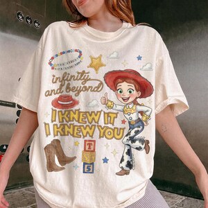 Może przedstawiać: Kremowy t-shirt z grafiką przedstawiającą kowbojkę, czerwony kapelusz, brązowe kowbojki i klocki z literami. Napis głosi "infinity and beyond" oraz "I KNEW IT I KNEW YOU", a całość uzupełnia grafika przedstawiająca naszyjnik z koralik&oacute;w.