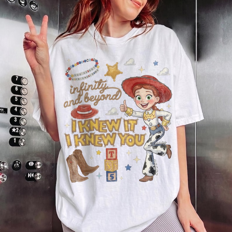 Może przedstawiać: Biały t-shirt z nadrukiem graficznym przedstawiającym kowbojkę, czerwony kapelusz kowbojski, brązowe kowbojki i stos klock&oacute;w z literami. Tekst głosi infinity and beyond, I KNEW IT oraz I KNEW YOU, wraz z grafiką kolorowego naszyjnika z koralik&oacute;w.