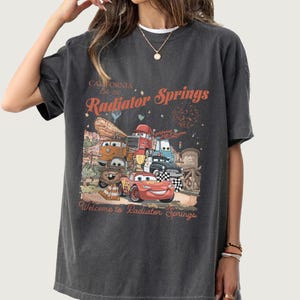 K&ouml;nnte beinhalten: Dunkelgraues T-Shirt mit einer Grafik von Charakteren aus dem Film Cars, darunter Lightning McQueen. Der Text auf dem Shirt lautet "California Est. 2006 Radiator Springs" und "Welcome to Radiator Springs."
