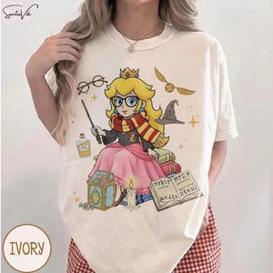 May include: Ivory-colored t-shirt featuring a cartoon princess with glasses, a wand, and a scarf, styled in a Harry Potter theme. The design includes a golden snitch, a sorting hat, and a potion bottle. The shirt has the word "IVORY" printed on it.