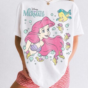 May include: A white t-shirt featuring a graphic print of Ariel from Disney's The Little Mermaid, along with Flounder and several seahorses surrounded by bubbles. The text reads Disney The Little Mermaid. The shirt is paired with red and white gingham shorts.