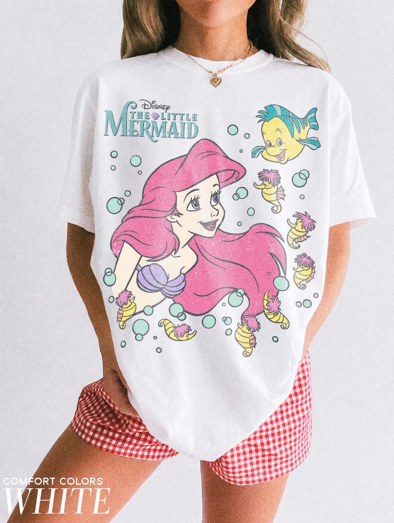May include: A white t-shirt featuring a graphic print of Ariel from Disney's The Little Mermaid, along with Flounder and several seahorses surrounded by bubbles. The text reads Disney The Little Mermaid. The shirt is paired with red and white gingham shorts.