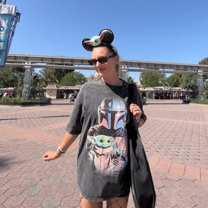 May include: A dark gray t-shirt featuring a graphic of The Mandalorian and Grogu. The shirt is worn with a black bag, sunglasses, and a headband with mouse ears and a Grogu design. The background shows a sunny outdoor setting.