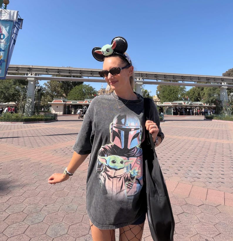 May include: A dark gray t-shirt featuring a graphic of The Mandalorian and Grogu. The shirt is worn with a black bag, sunglasses, and a headband with mouse ears and a Grogu design. The background shows a sunny outdoor setting.