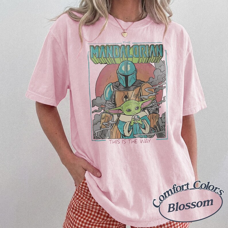 May include: A light pink short-sleeve t-shirt featuring a graphic print of The Mandalorian and Grogu. The design includes the text THE MANDALORIAN and THIS IS THE WAY, set against a circular background with smoke elements and a rectangular border.