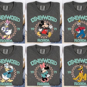 May include: Six gray t-shirts featuring Disney characters. Each shirt has the words "Disney World" in teal, the year "1971", and "Florida". The characters include Daisy Duck, Mickey Mouse, Goofy, Donald Duck, Minnie Mouse, and Pluto.