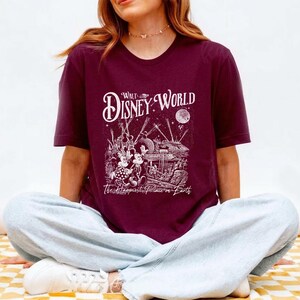 May include: Burgundy t-shirt featuring a white graphic of the Walt Disney World logo and a detailed illustration of the park with Mickey and Minnie Mouse. The text "The Happiest Place on Earth" is below the illustration.