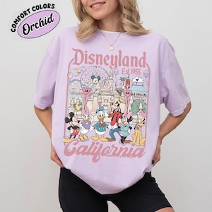 May include: A light purple t-shirt featuring a Disneyland graphic with Mickey Mouse, Donald Duck, and other cartoon characters. The graphic includes the words "Disneyland California Est. 1955" in pink lettering. The shirt has a relaxed fit.