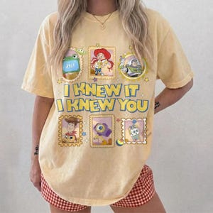 May include: A pale yellow short-sleeve t-shirt featuring a graphic print of Toy Story characters in framed boxes. The text "I KNEW IT I KNEW YOU" is printed in large blue letters in the center, surrounded by small stars and colorful icons.