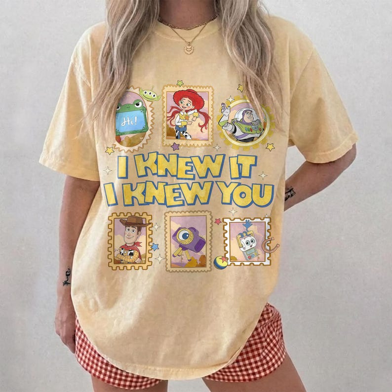 May include: A pale yellow short-sleeve t-shirt featuring a graphic print of Toy Story characters in framed boxes. The text "I KNEW IT I KNEW YOU" is printed in large blue letters in the center, surrounded by small stars and colorful icons.