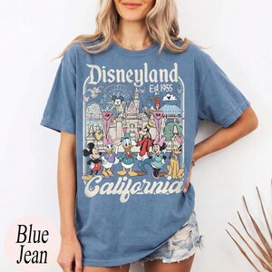 May include: Blue jean t-shirt featuring a Disneyland graphic with Mickey Mouse, Minnie Mouse, Donald Duck, Goofy, and other characters. The graphic includes the text "Disneyland California Est. 1955". The shirt is a light blue color.