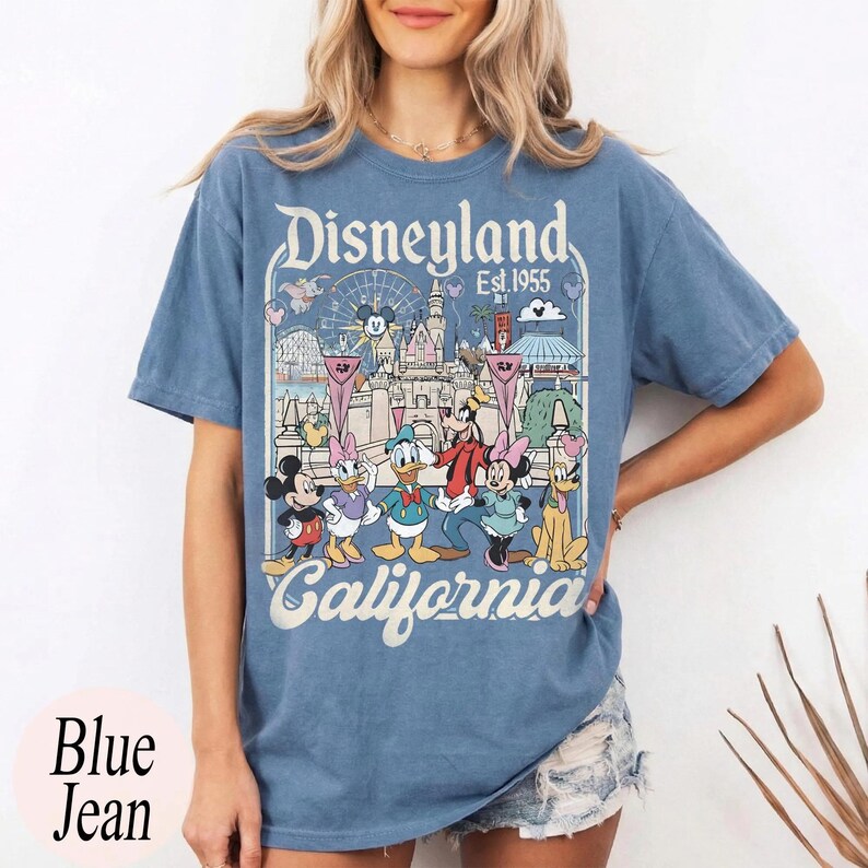 May include: Blue jean t-shirt featuring a Disneyland graphic with Mickey Mouse, Minnie Mouse, Donald Duck, Goofy, and other characters. The graphic includes the text "Disneyland California Est. 1955". The shirt is a light blue color.