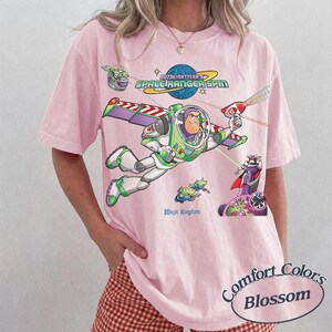 May include: A light pink short-sleeve t-shirt featuring a graphic print of Buzz Lightyear from Space Ranger Spin. The design includes the text Buzz Lightyear's Space Ranger Spin and Magic Kingdom, with colorful illustrations of characters.
