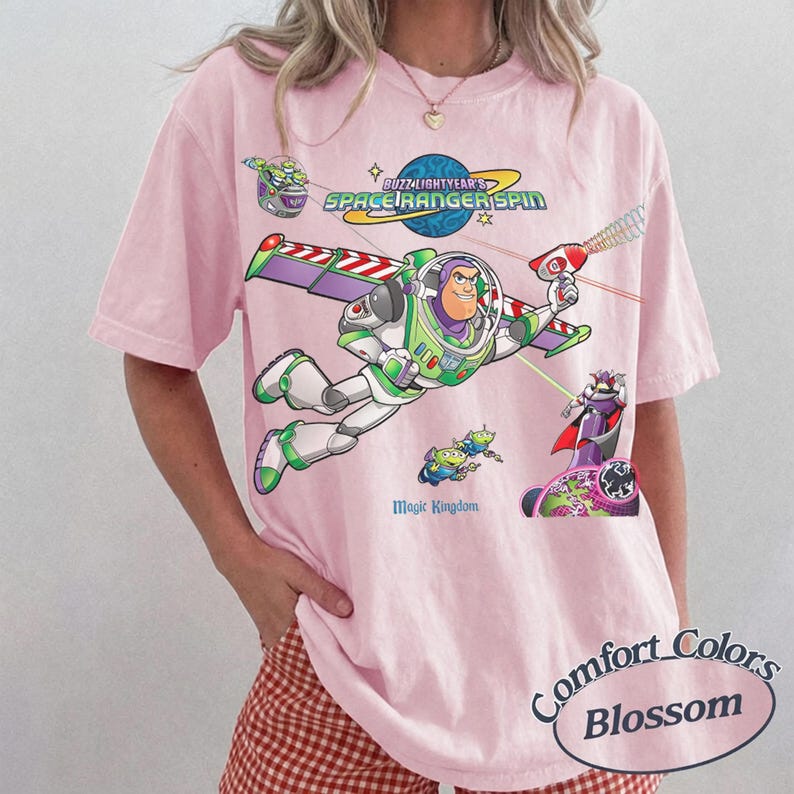 May include: A light pink short-sleeve t-shirt featuring a graphic print of Buzz Lightyear from Space Ranger Spin. The design includes the text Buzz Lightyear's Space Ranger Spin and Magic Kingdom, with colorful illustrations of characters.