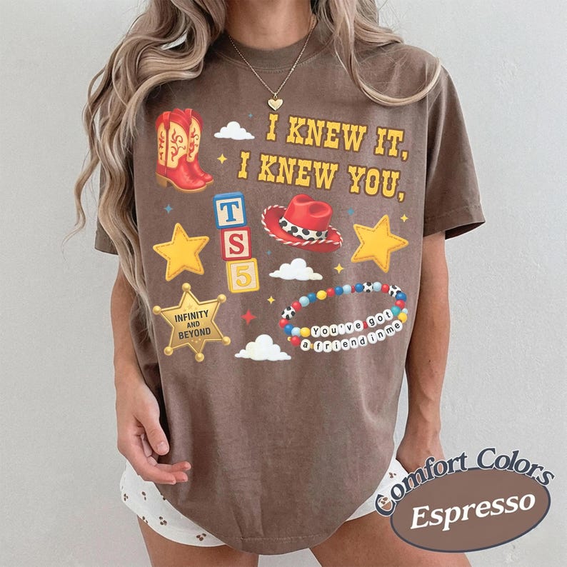 May include: A brown short-sleeve t-shirt featuring yellow text that reads I KNEW IT, I KNEW YOU, along with graphics of a red cowboy boot, a red cowboy hat, a sheriff badge with the text INFINITY AND BEYOND, alphabet blocks, and a beaded bracelet.