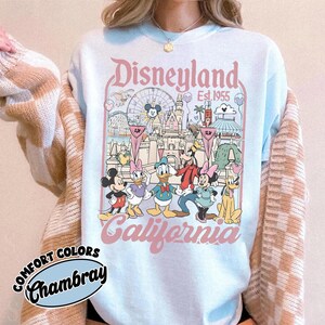 May include: Light blue sweatshirt featuring a Disneyland graphic with Mickey Mouse, Minnie Mouse, Donald Duck, Goofy, and Daisy Duck. The graphic includes the text "Disneyland California Est. 1955". The sweatshirt has the text "Comfort Colors Chambray" on the bottom left.