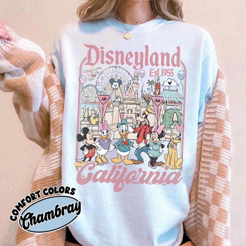 May include: Light blue sweatshirt featuring a Disneyland graphic with Mickey Mouse, Minnie Mouse, Donald Duck, Goofy, and Daisy Duck. The graphic includes the text "Disneyland California Est. 1955". The sweatshirt has the text "Comfort Colors Chambray" on the bottom left.