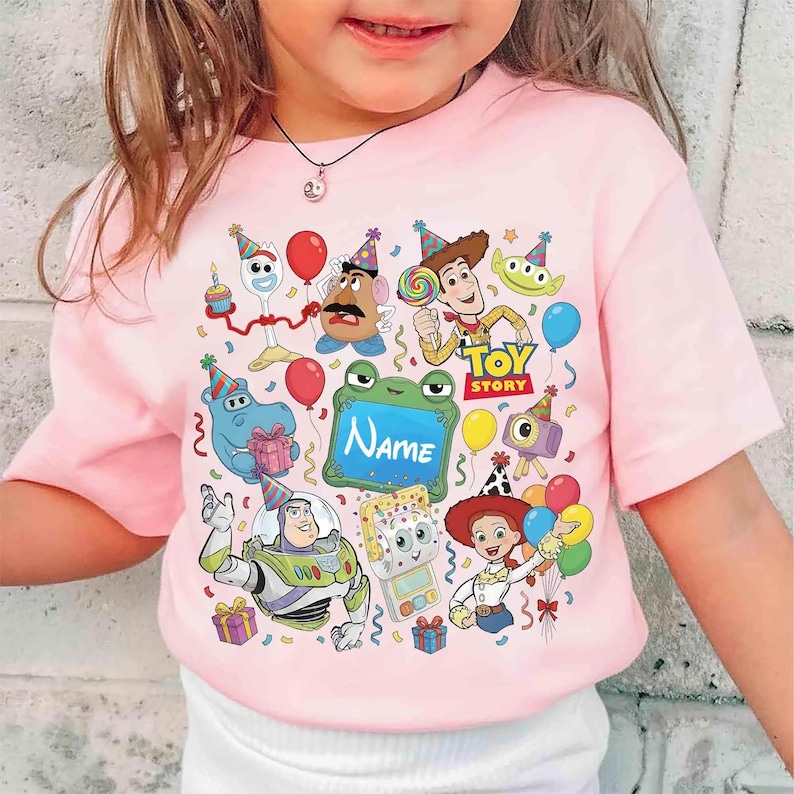 May include: A light pink short-sleeved t-shirt featuring a colorful graphic print of various Toy Story characters including Woody, Buzz Lightyear, Jessie, and Forky, along with balloons, party hats, and a central green frame with the text Name.