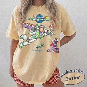 May include: A yellow short-sleeve t-shirt featuring a graphic of Buzz Lightyear flying with the text Buzz Lightyear Space Ranger Spin and Magic Kingdom. The design includes a small green alien and a purple character on a circular base with a laser beam.